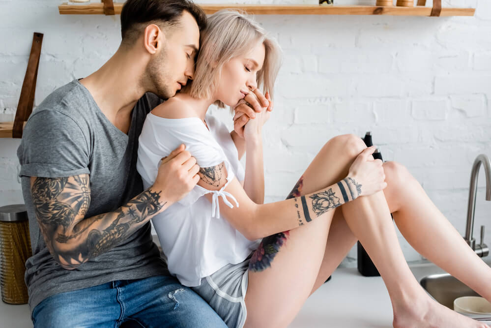 Tattooed man embracing beautiful blonde girlfriend on kitchen worktop