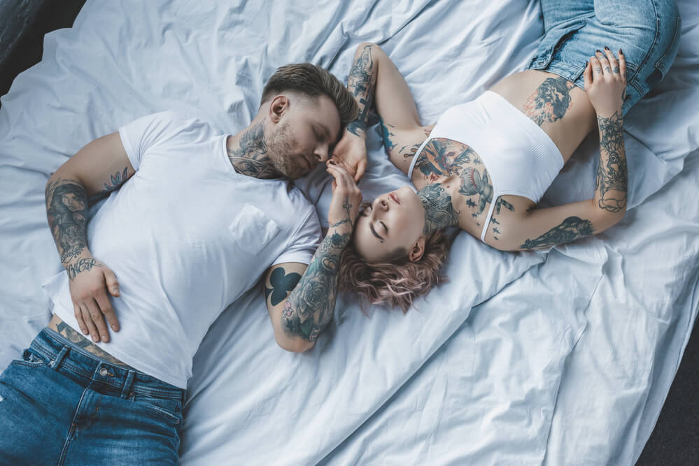 Top view of tender tattooed lovers holding hands and relaxing on white bad