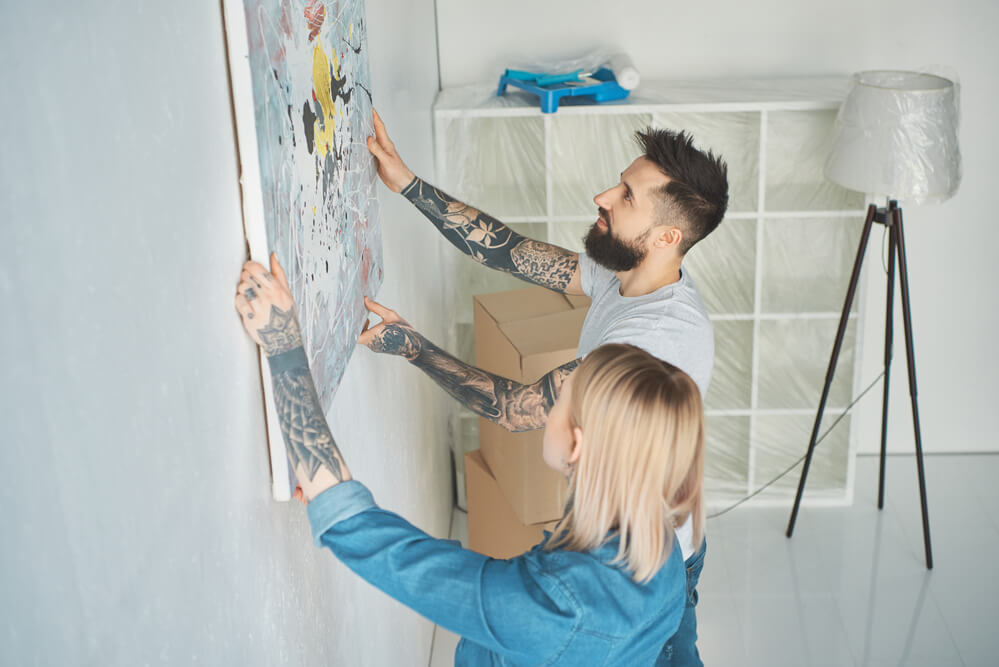 Side view of young tattooed couple hanging painting on wall at new apartment