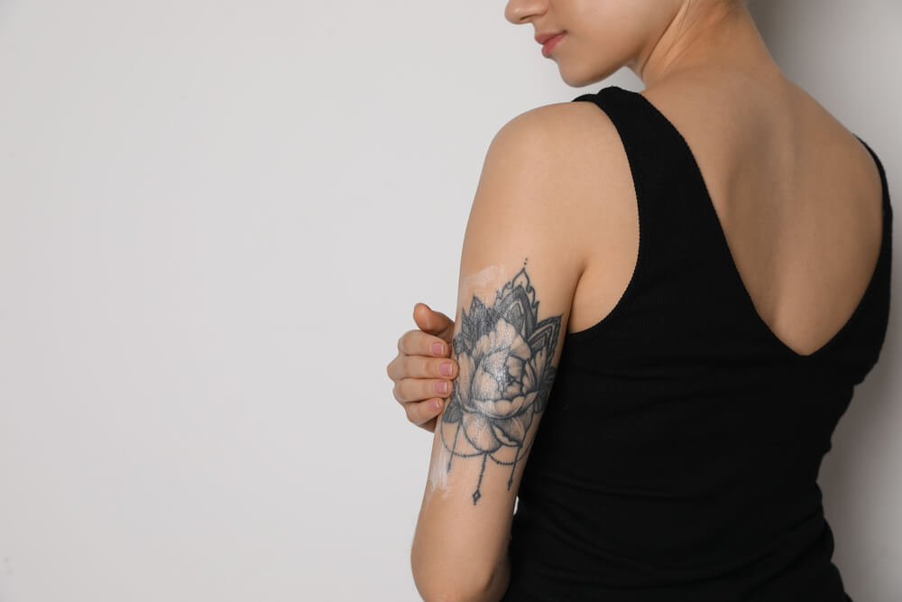 Woman applying cream onto her arm with tattoo on white background, closeup. Space for text